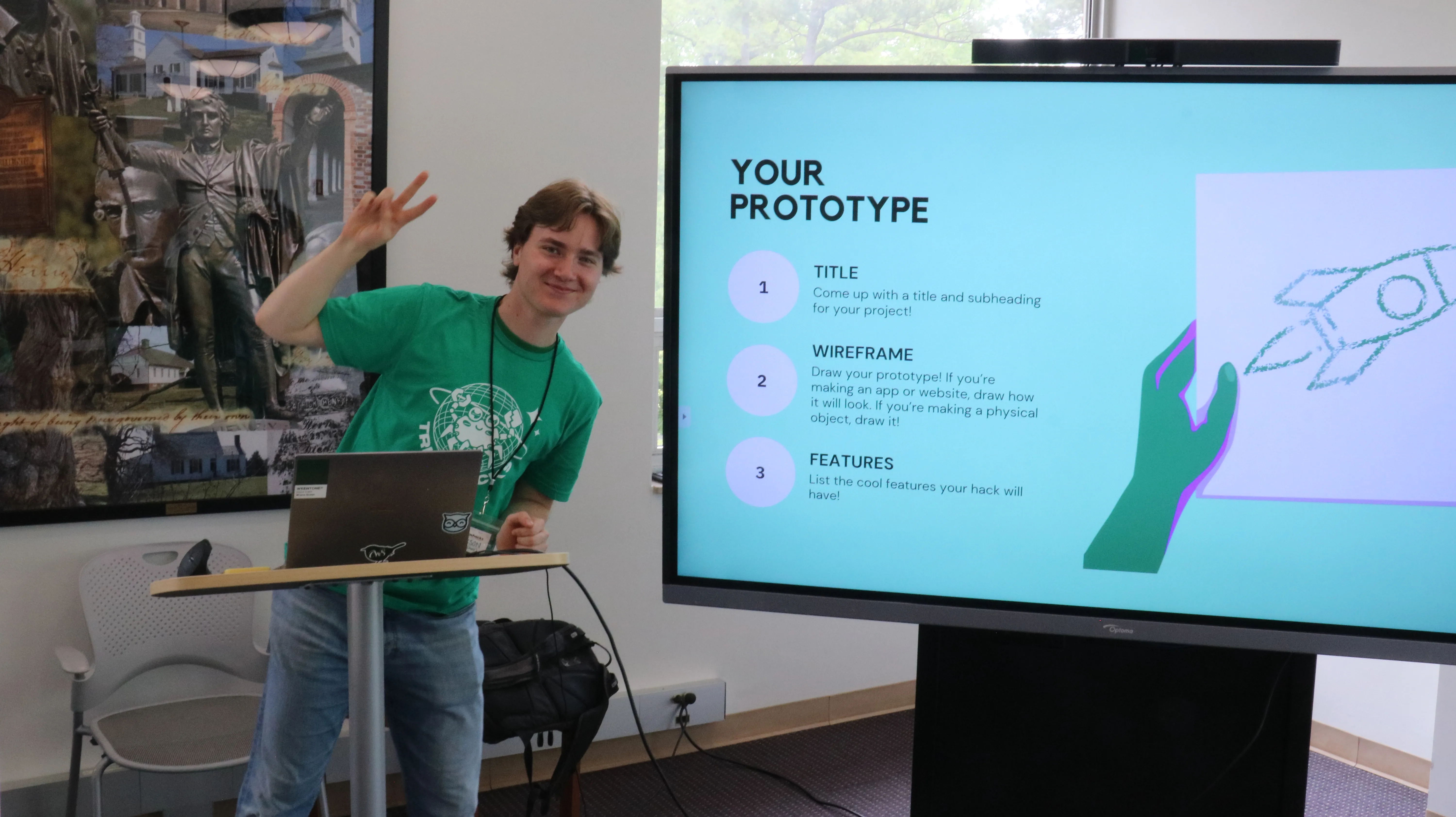 A person leaning to the side, holding up two fingers in a peace sign. They are standing in front of a whiteboard that says 'Your Prototype'